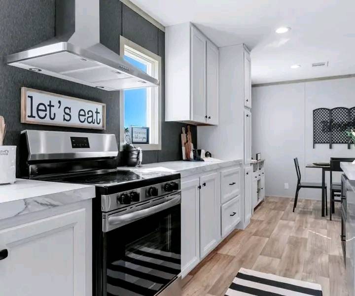 A modern kitchen with a stainless steel oven and hood, white cabinets, and marble countertops. A sign reads "let's eat." Light wood flooring adds warmth.