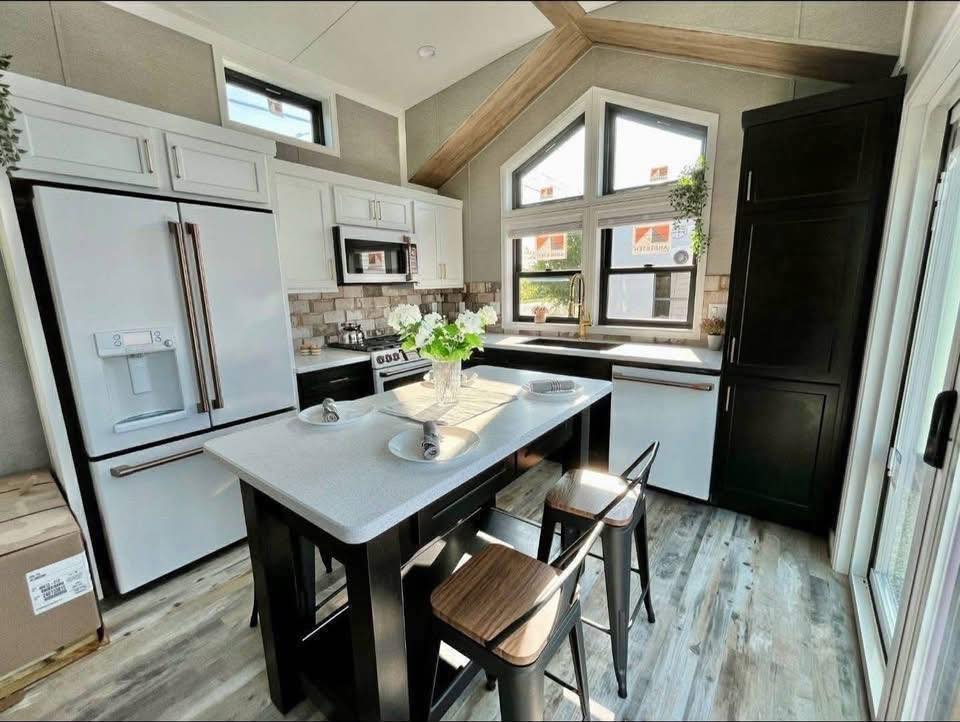 Modern kitchen with a high ceiling and large triangular window. Features white cabinets, stainless steel appliances, a dark island, and light wood flooring.