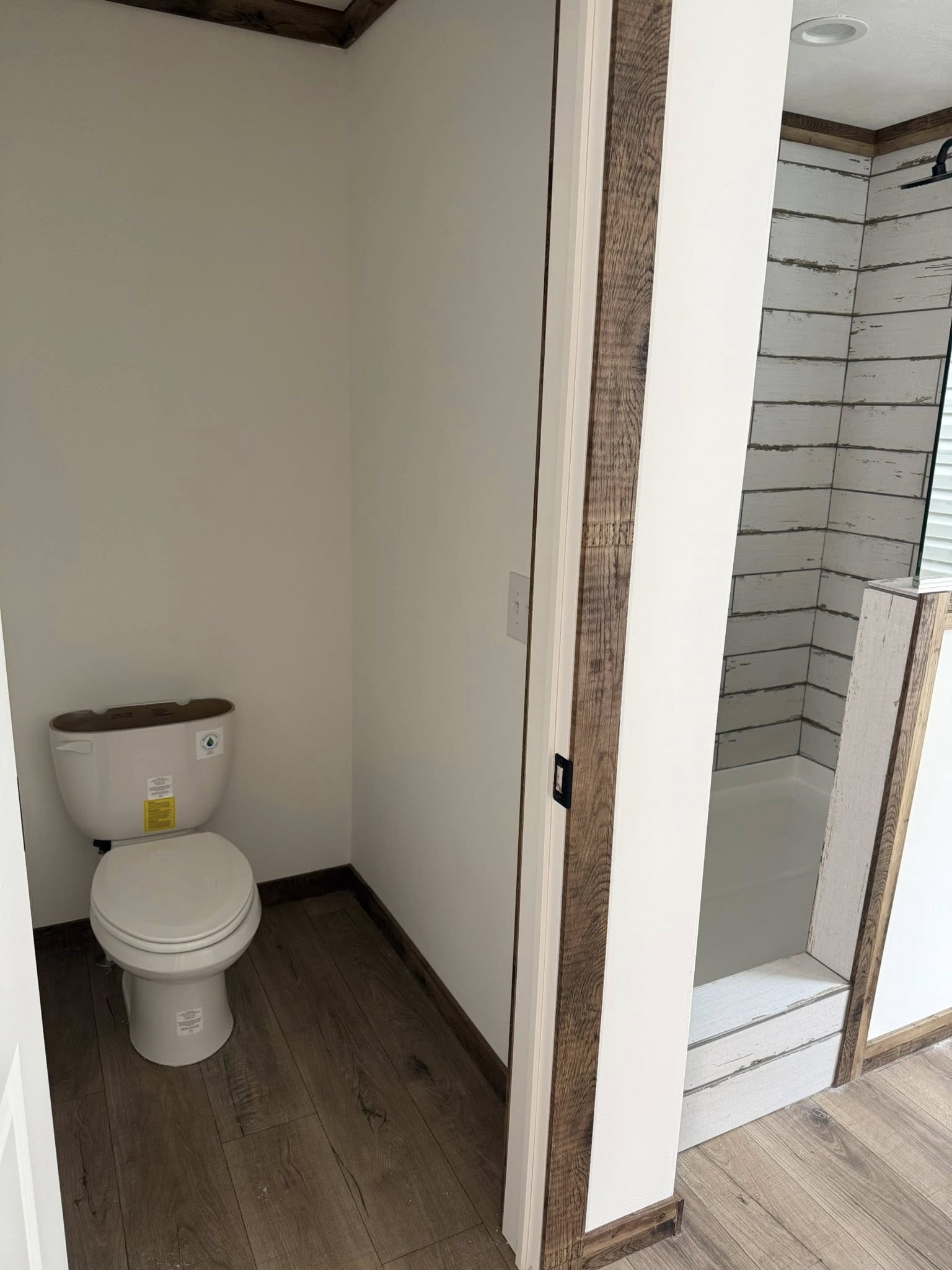 A small bathroom with a white toilet on a wooden floor. Adjacent is a shower with rustic, whitewashed wood paneling. The atmosphere is minimalist.