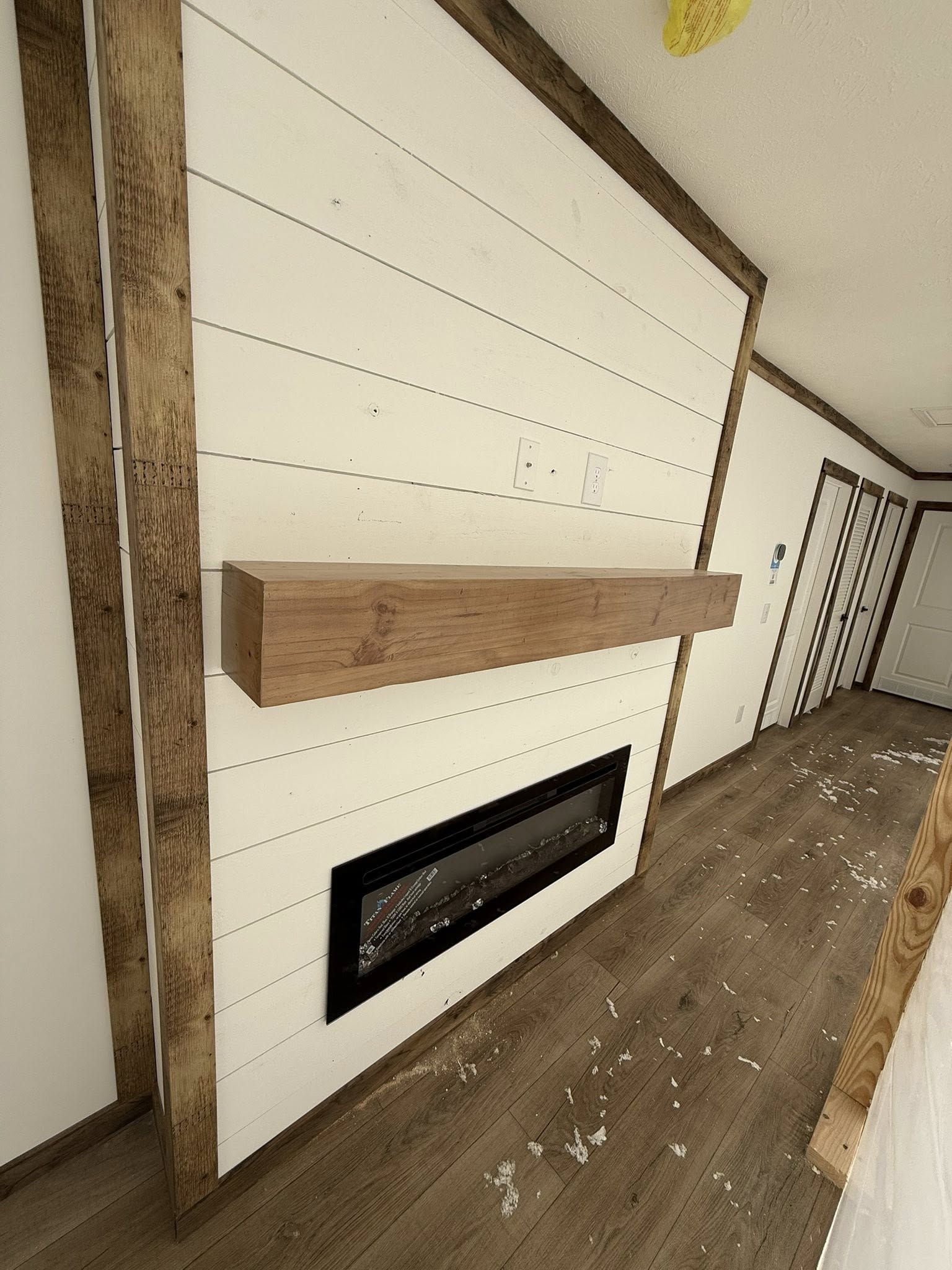 Wall with white horizontal shiplap, featuring a rustic wooden beam shelf and an inset black electric fireplace. The wood floor has debris scattered.