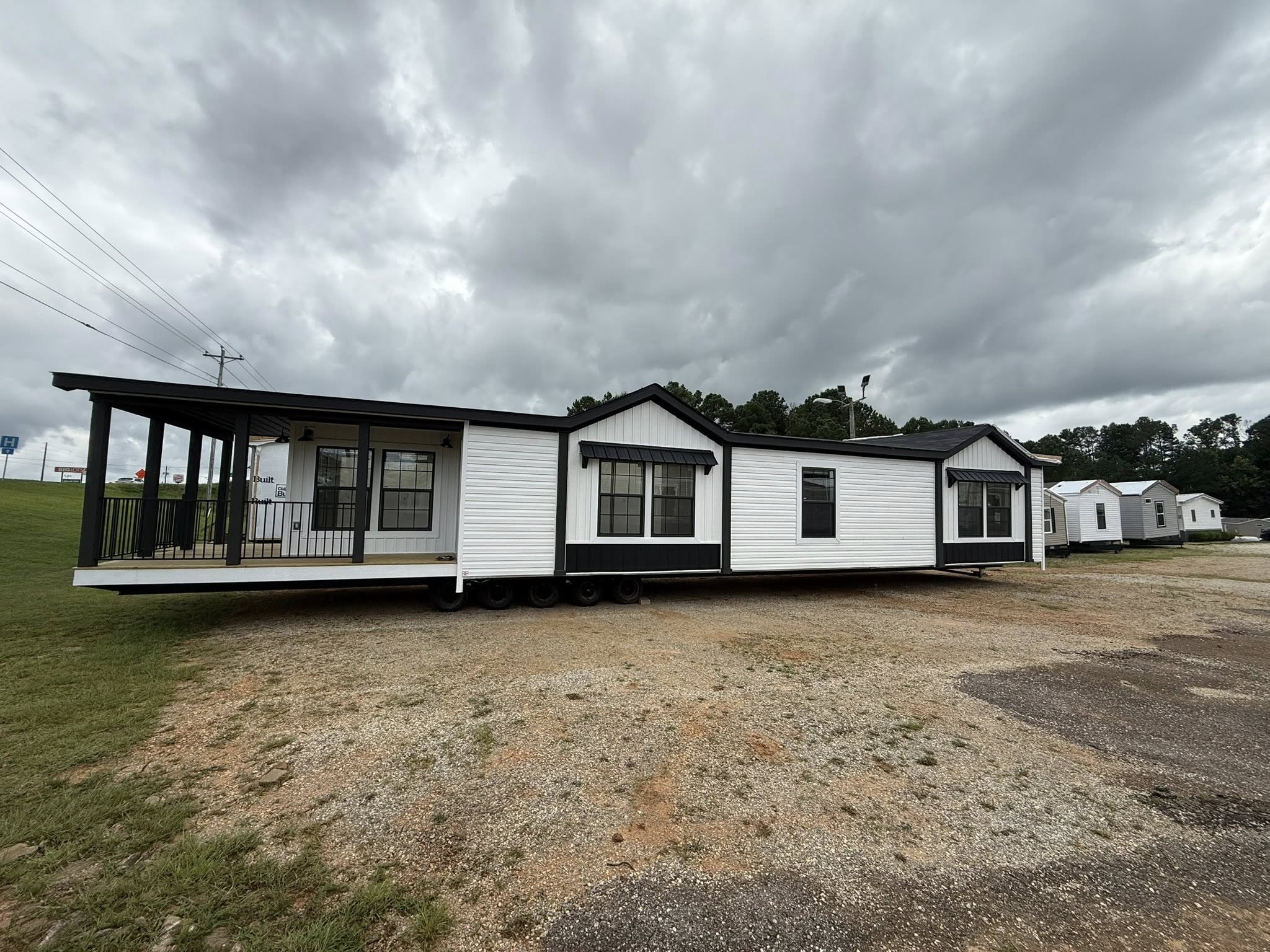 Large white and black mobile home on a lot, under a cloudy sky. Features a porch with railings and multiple windows. Overcast and subdued atmosphere.