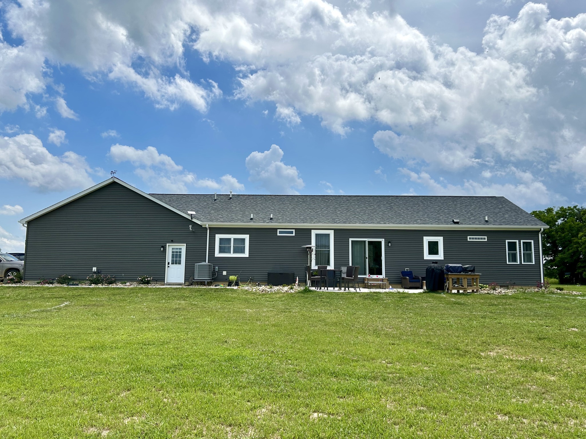 Single-story gray house with white doors and large windows, set against a bright blue sky with fluffy clouds. A lush green lawn surrounds the home.