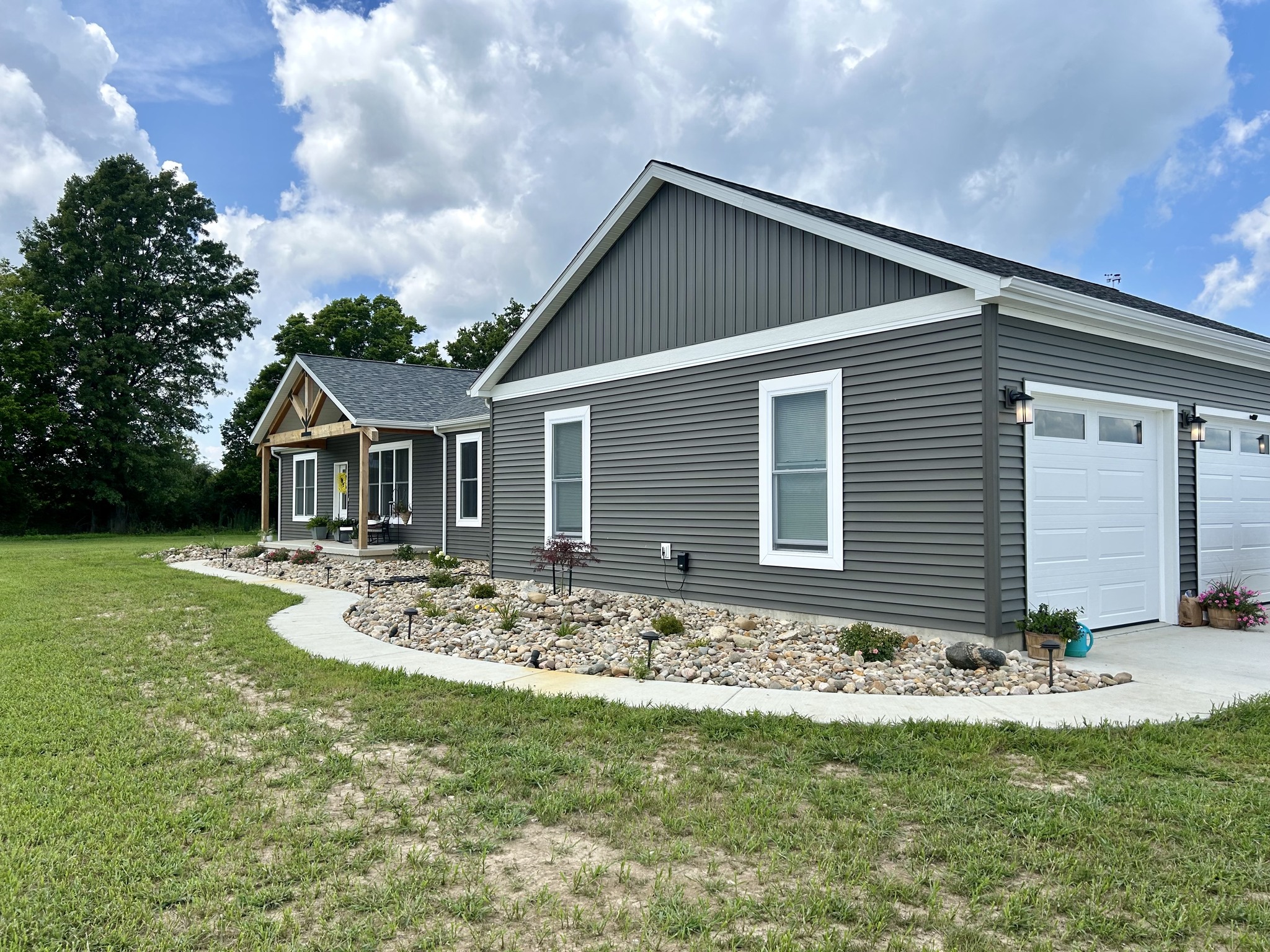Modern gray house with white trim, surrounded by a stone garden and lush green lawn. Bright sky with fluffy clouds overhead, creating a peaceful atmosphere.