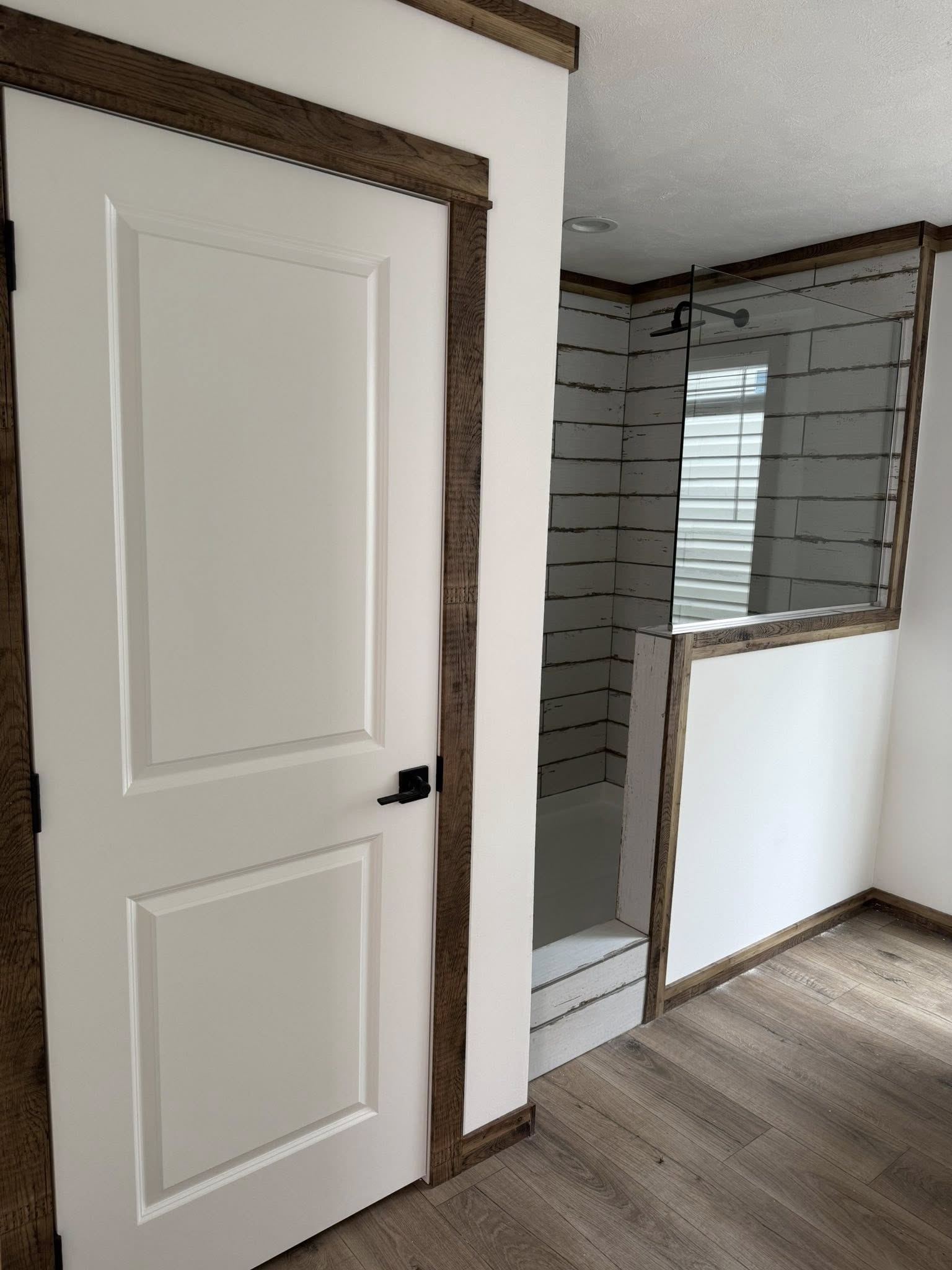A bathroom with a modern look, featuring a door with wooden trim, a glass shower enclosure with white wood-patterned tiles, and wood flooring.