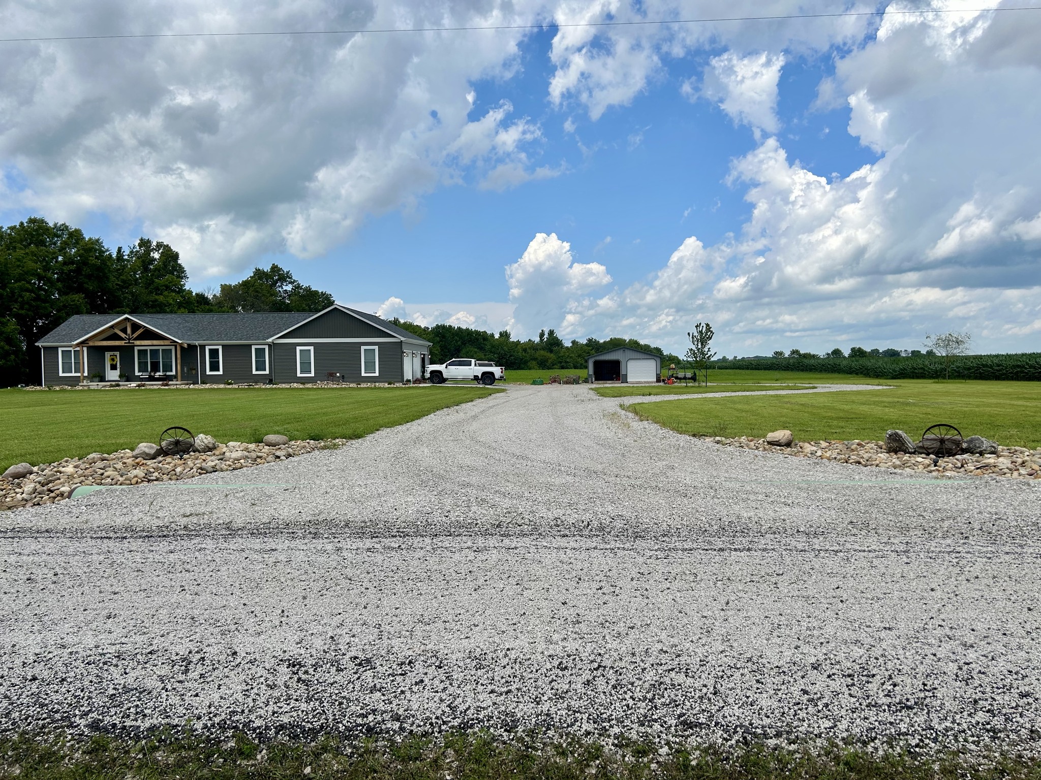 A gravel driveway leads to a gray house with a porch, surrounded by green lawn and trees under a partly cloudy sky, evoking a peaceful, rural vibe.