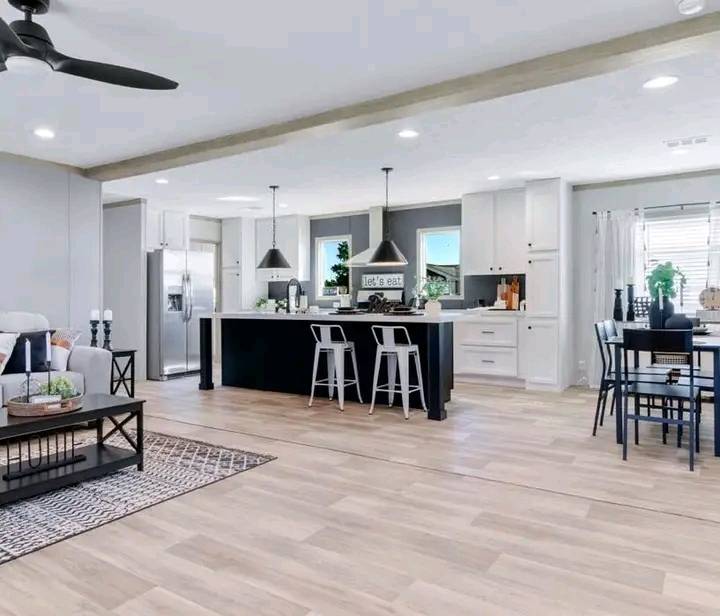 Modern open-plan kitchen and living area with wooden flooring. Features a sleek island with stools, pendant lights, a dining table, and cozy seating. Bright, airy ambiance.