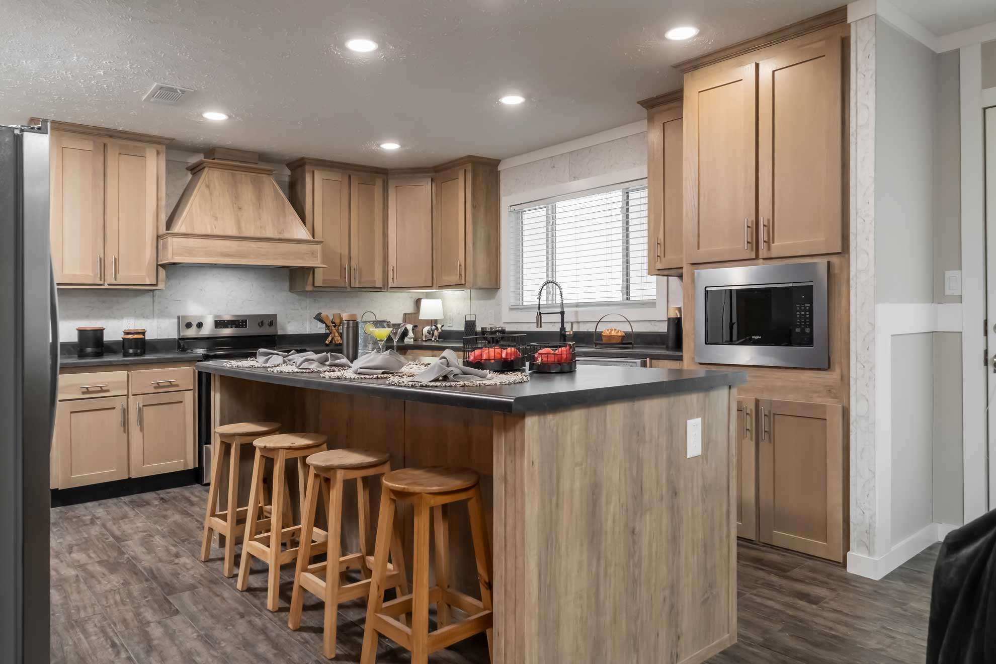 Modern kitchen with wood cabinets, island, and four wooden stools. Stainless steel appliances, large window, under soft lighting, creating a warm ambiance.