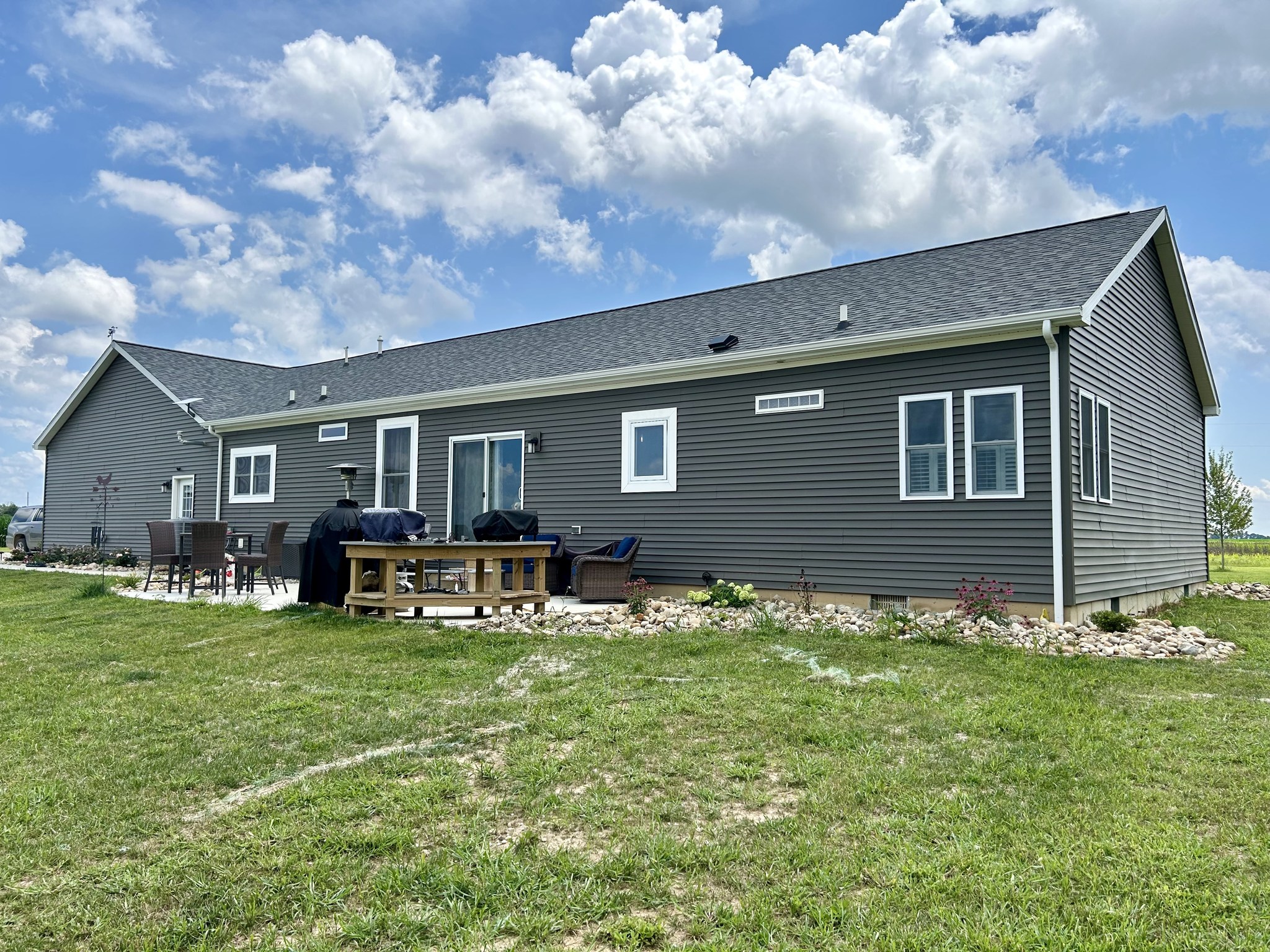Single-story gray house with white trim, patio furniture, and a barbecue in a backyard with green grass and a bright blue sky with fluffy clouds.