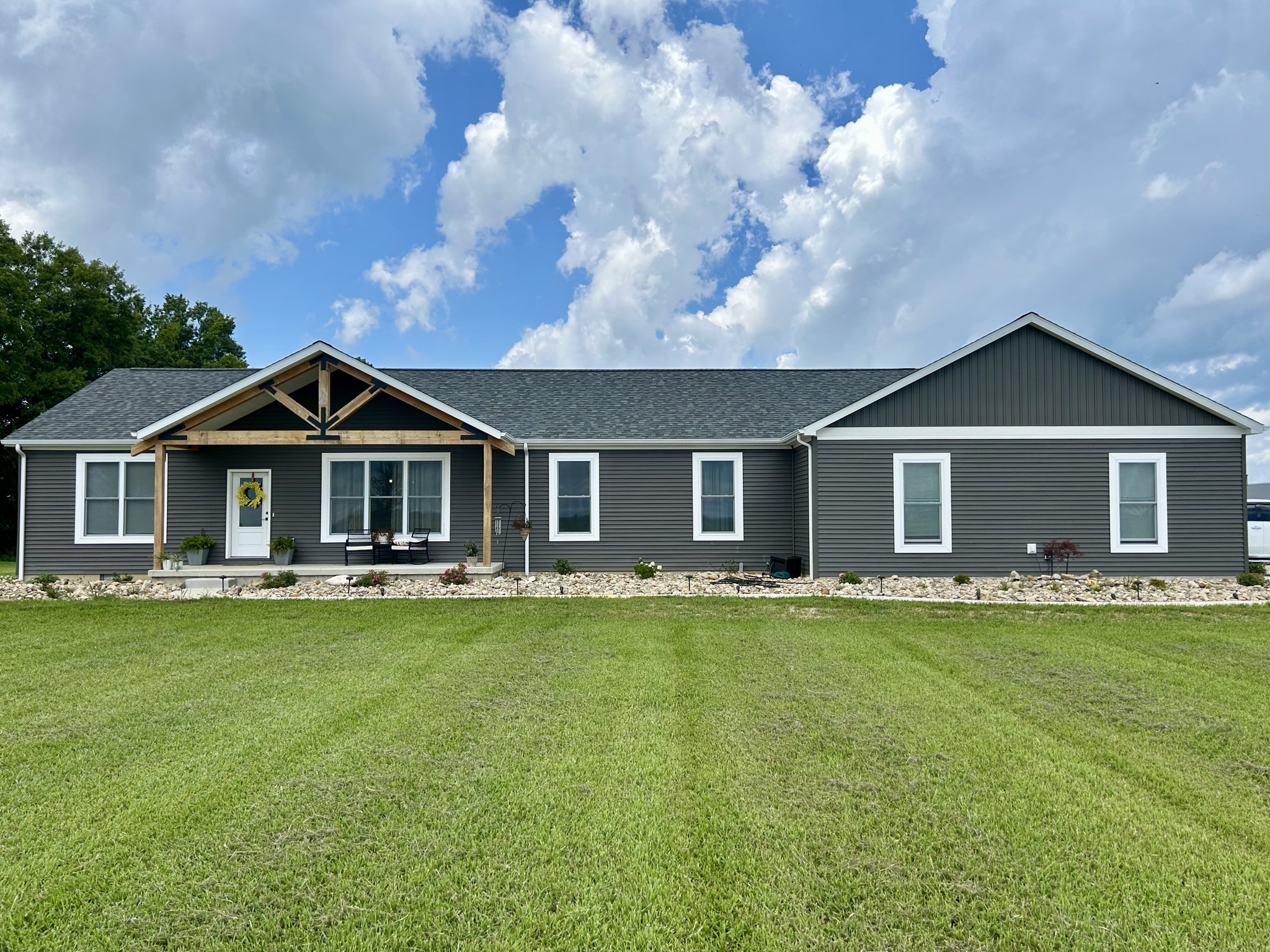 Single-story gray house with a gabled roof, white trim, and a porch. Set against a vibrant green lawn and a cloudy blue sky. Peaceful atmosphere.