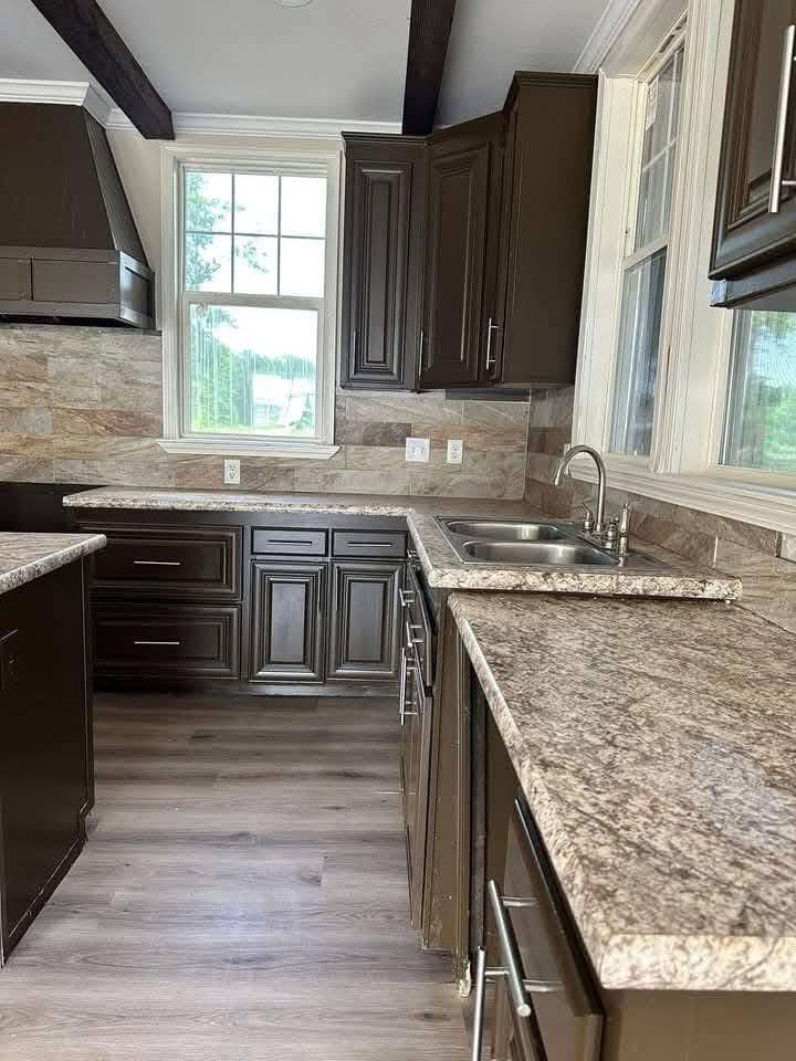Modern kitchen with dark cabinets, marble countertops, and wood flooring. Large windows provide natural light, creating a warm, inviting atmosphere.