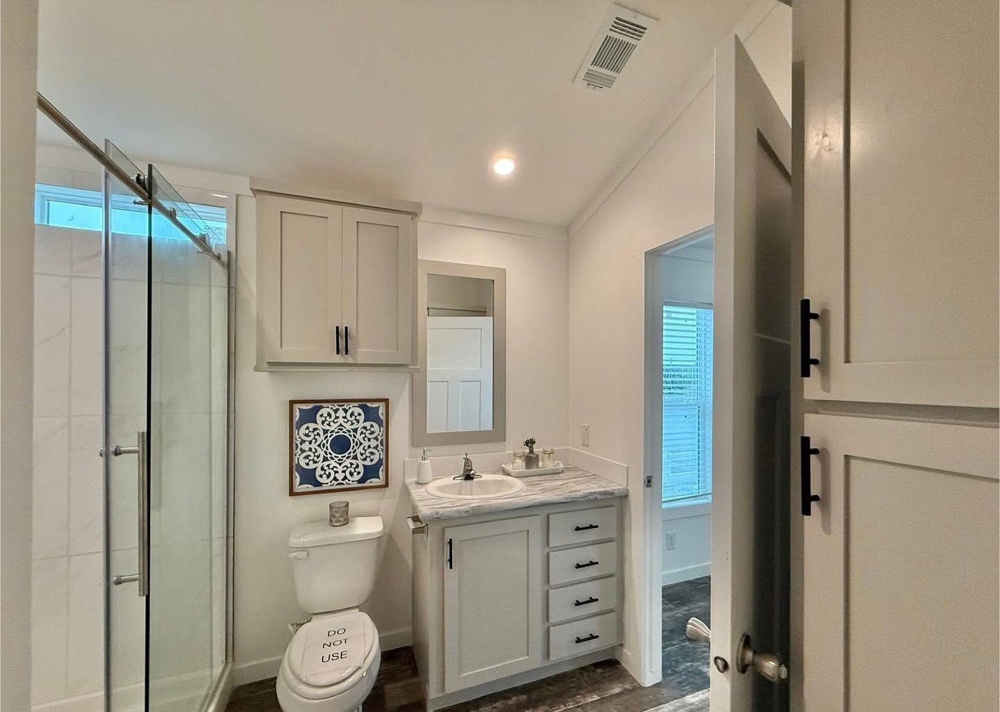 A modern bathroom features a glass shower, a vanity with a marble countertop, and a toilet with a "Do Not Use" sign. Bright and clean design.