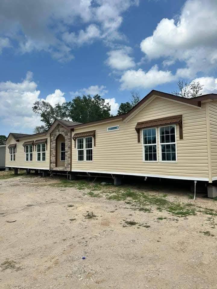 A beige manufactured home with a stone entryway sits on a dirt lot under a partly cloudy sky, conveying a sense of simplicity and tranquility.