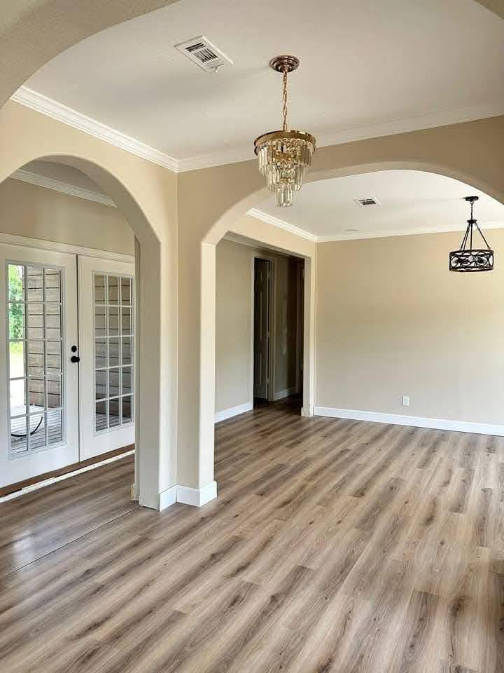 Bright, empty room with wood flooring, cream walls, and two chandeliers. Arched entryways lead to a sunlit area with French doors. Airy and elegant.