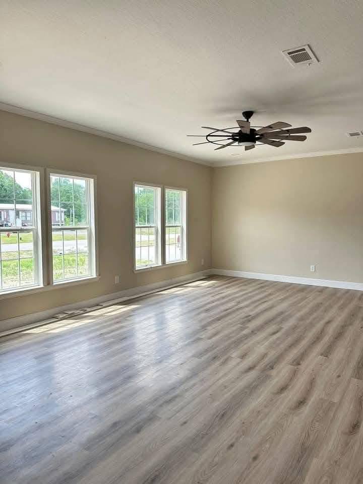 A spacious, empty room with light wood flooring, beige walls, and a ceiling fan. Large windows let in natural light, creating a bright and airy feel.