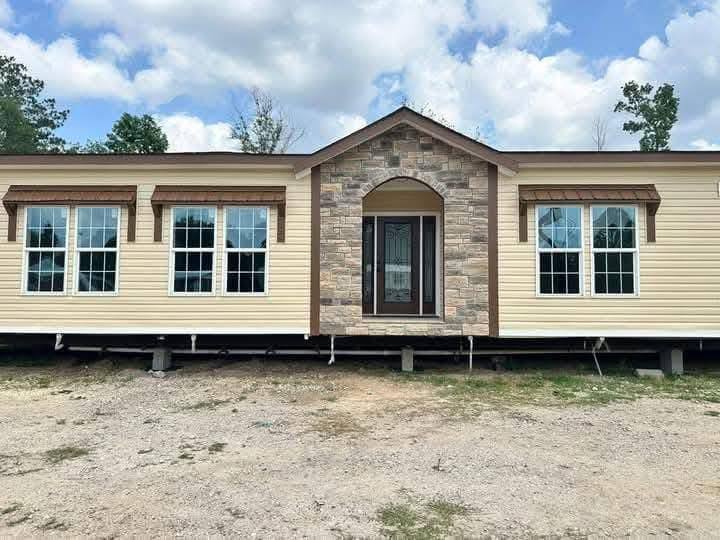 A beige modular home with stone accents is elevated on blocks. It features large windows and a central door under a partly cloudy sky.