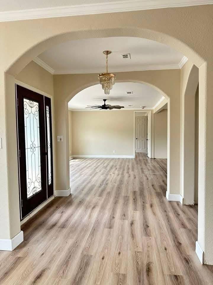 A spacious hallway with light wood flooring, beige walls, and decorative archways. A chandelier hangs above, leading to a room with a ceiling fan.