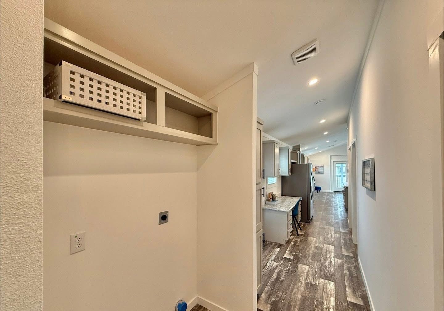 Hallway view leading to a modern kitchen with wood-patterned flooring. Shelving with a white basket is on the left, evoking a neat, minimalist atmosphere.