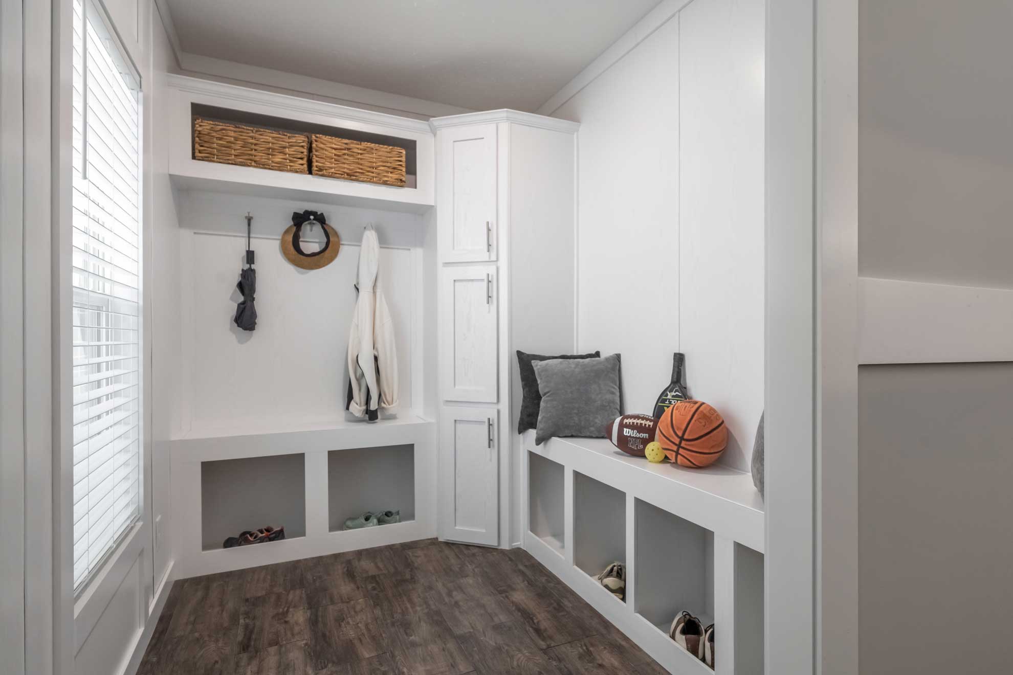 Modern mudroom with white cabinets and cubbies. Baskets, coats, and shoes are neatly organized. Sports equipment and cushions add a cozy touch.