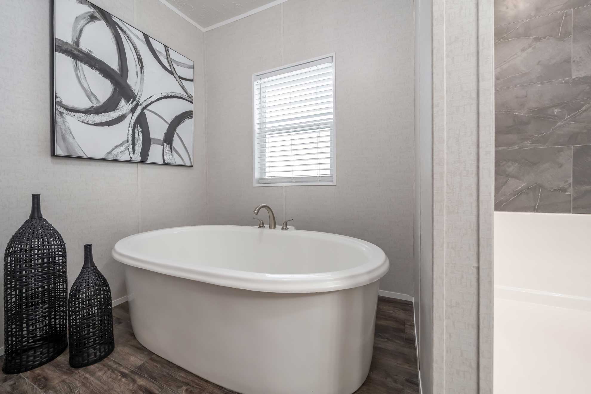 Minimalist bathroom featuring a white freestanding bathtub, abstract black-and-white art, large wicker vases, and a window with blinds, conveying tranquility.