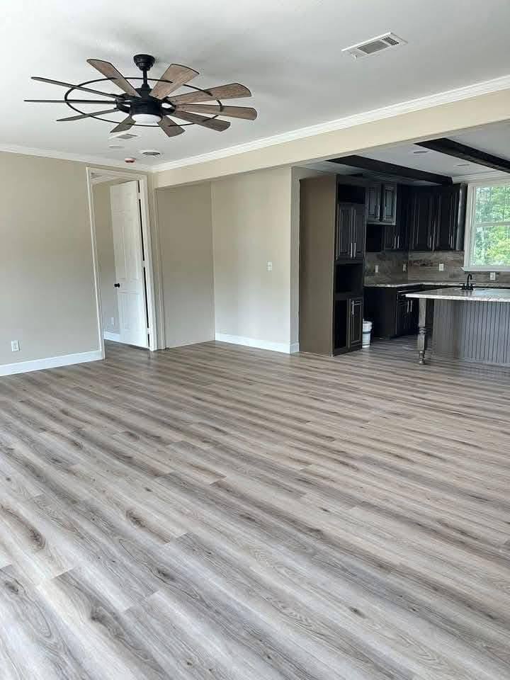 Spacious open-plan room with light wood flooring, a large modern ceiling fan, and a kitchen with dark cabinets. Bright, airy atmosphere.