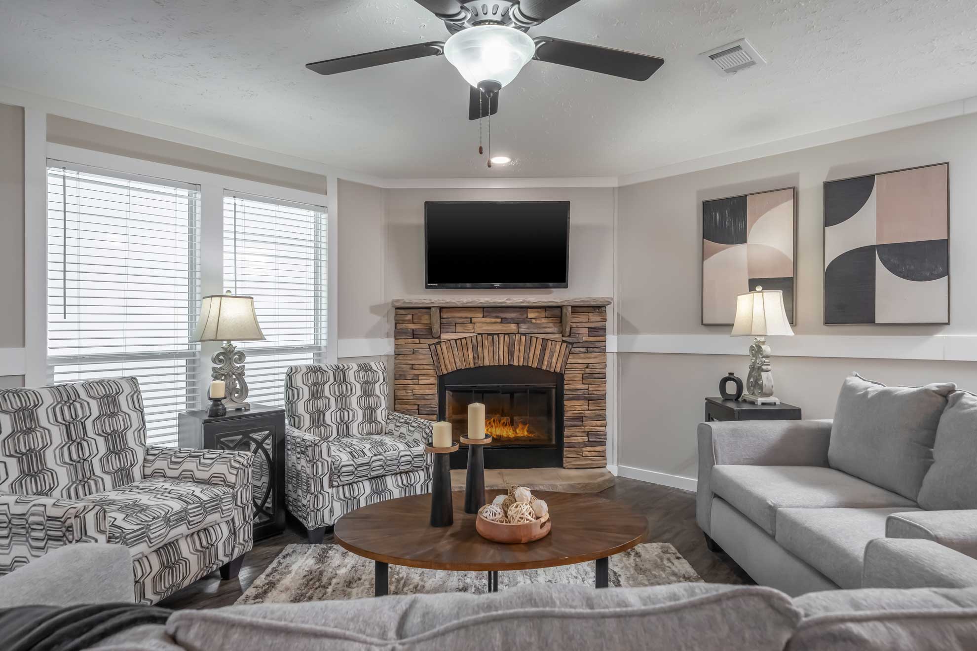 Modern living room with a cozy stone fireplace, two patterned armchairs, gray sofa, abstract wall art, ceiling fan, and warm lighting from lamps.