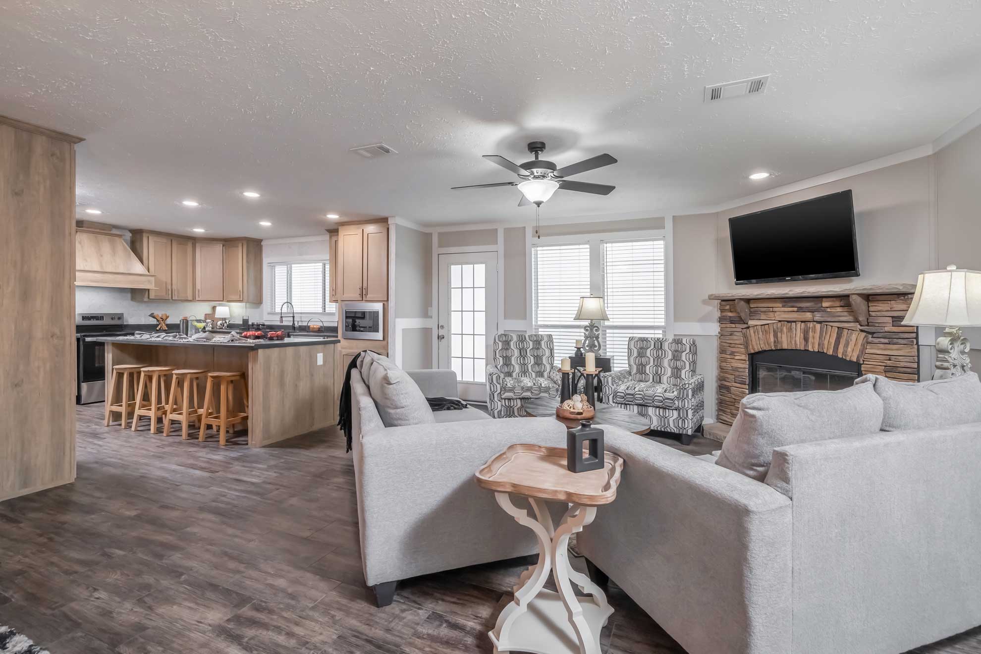 Bright open-concept living room and kitchen with gray sofas, stone fireplace, ceiling fan, and wide windows. Kitchen has wooden cabinets and island. Cozy and inviting ambiance.