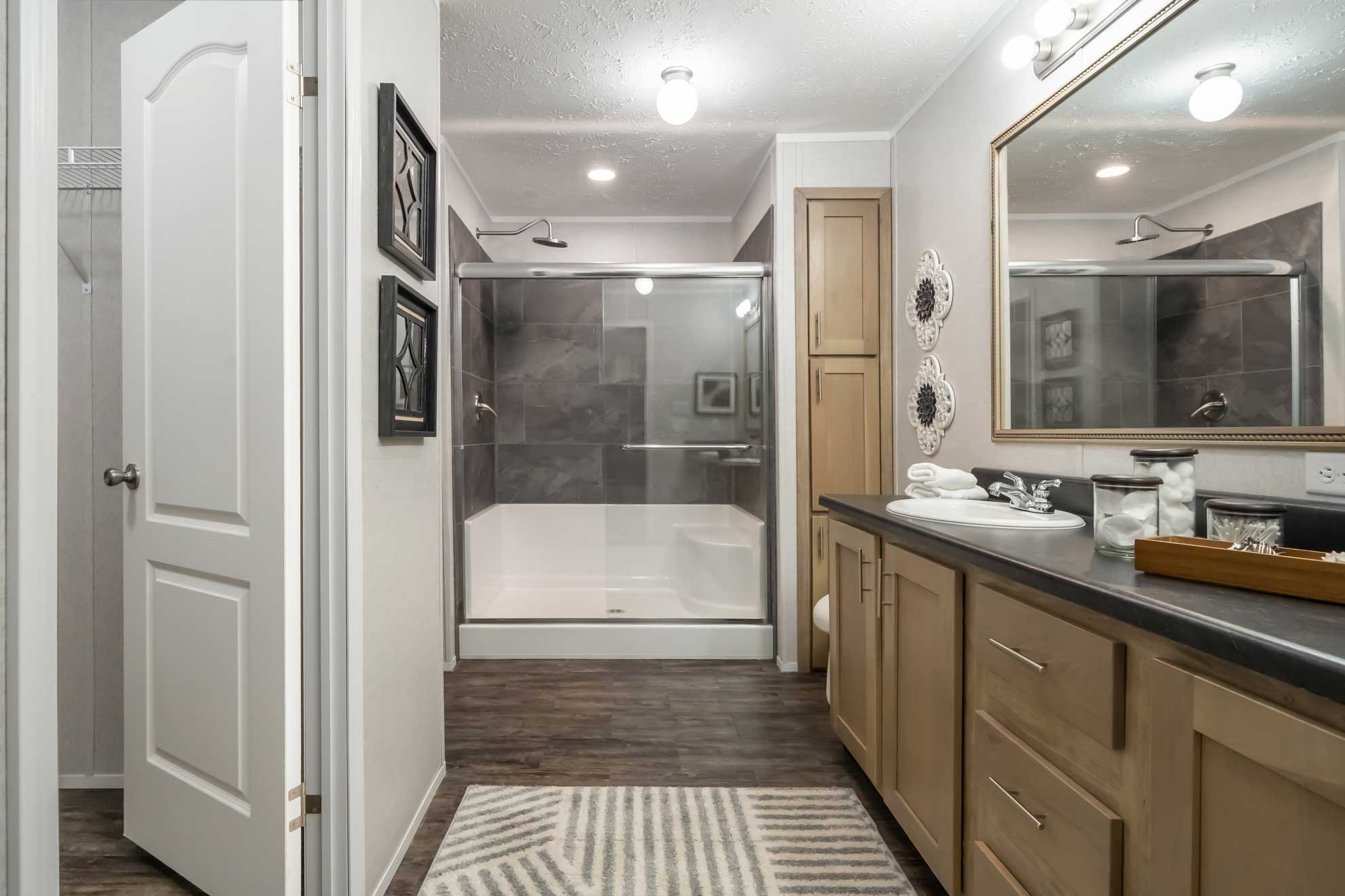 Modern bathroom with wood cabinets, large mirror, and marble countertop. Glass shower with gray tiles, white bathtub, and a striped rug on wooden floor.