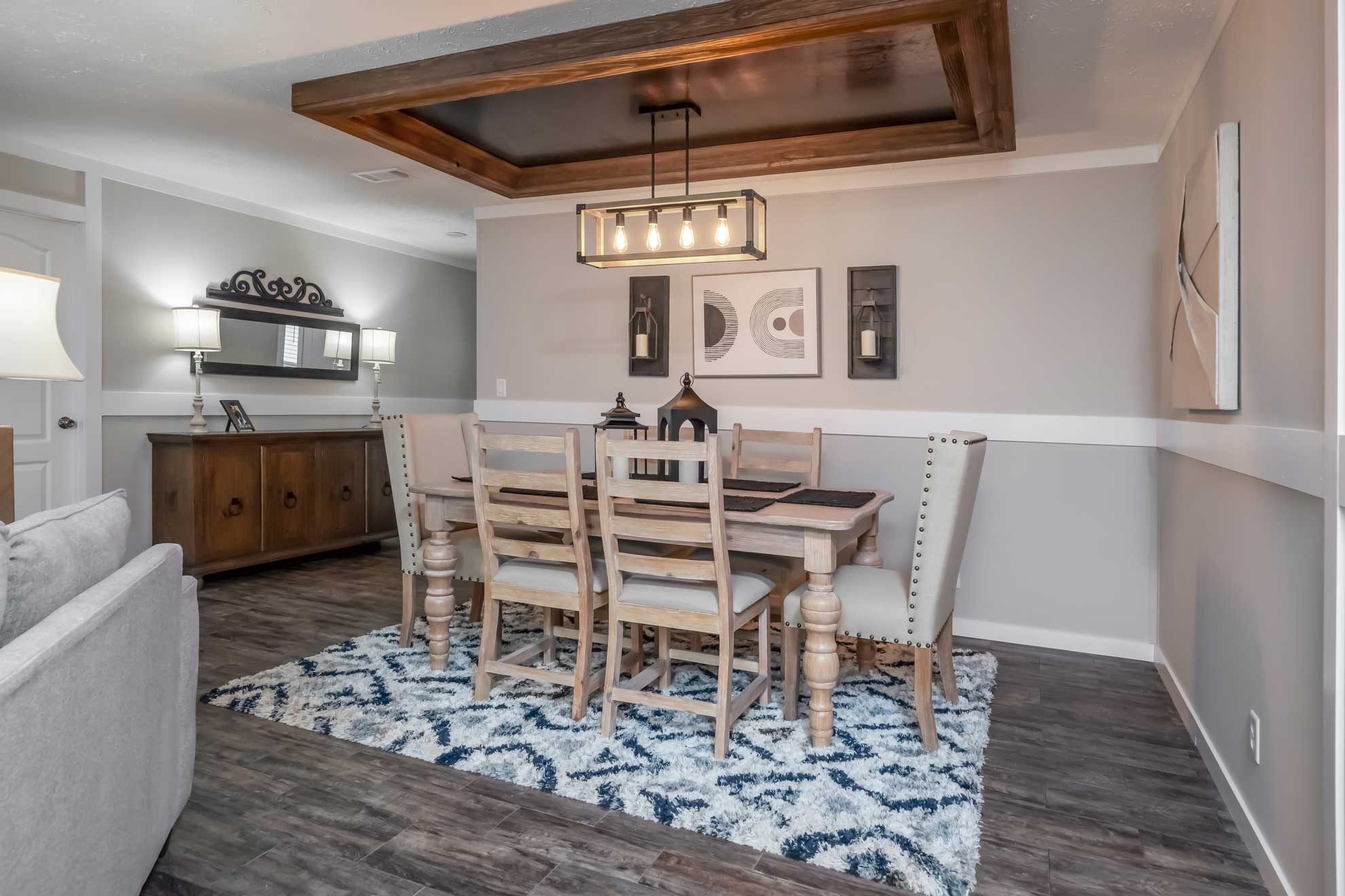 Dining room with a wooden table and six upholstered chairs on a patterned rug. A modern chandelier hangs above. Artwork and a sideboard enhance the cozy ambiance.