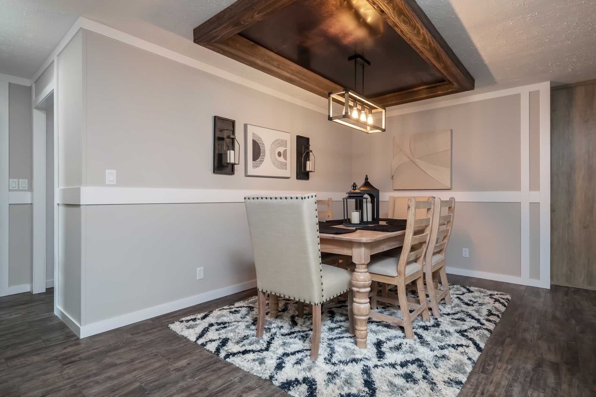 Dining room with a wooden table and six chairs on a patterned rug. Modern pendant light and abstract wall art create a cozy, elegant atmosphere.
