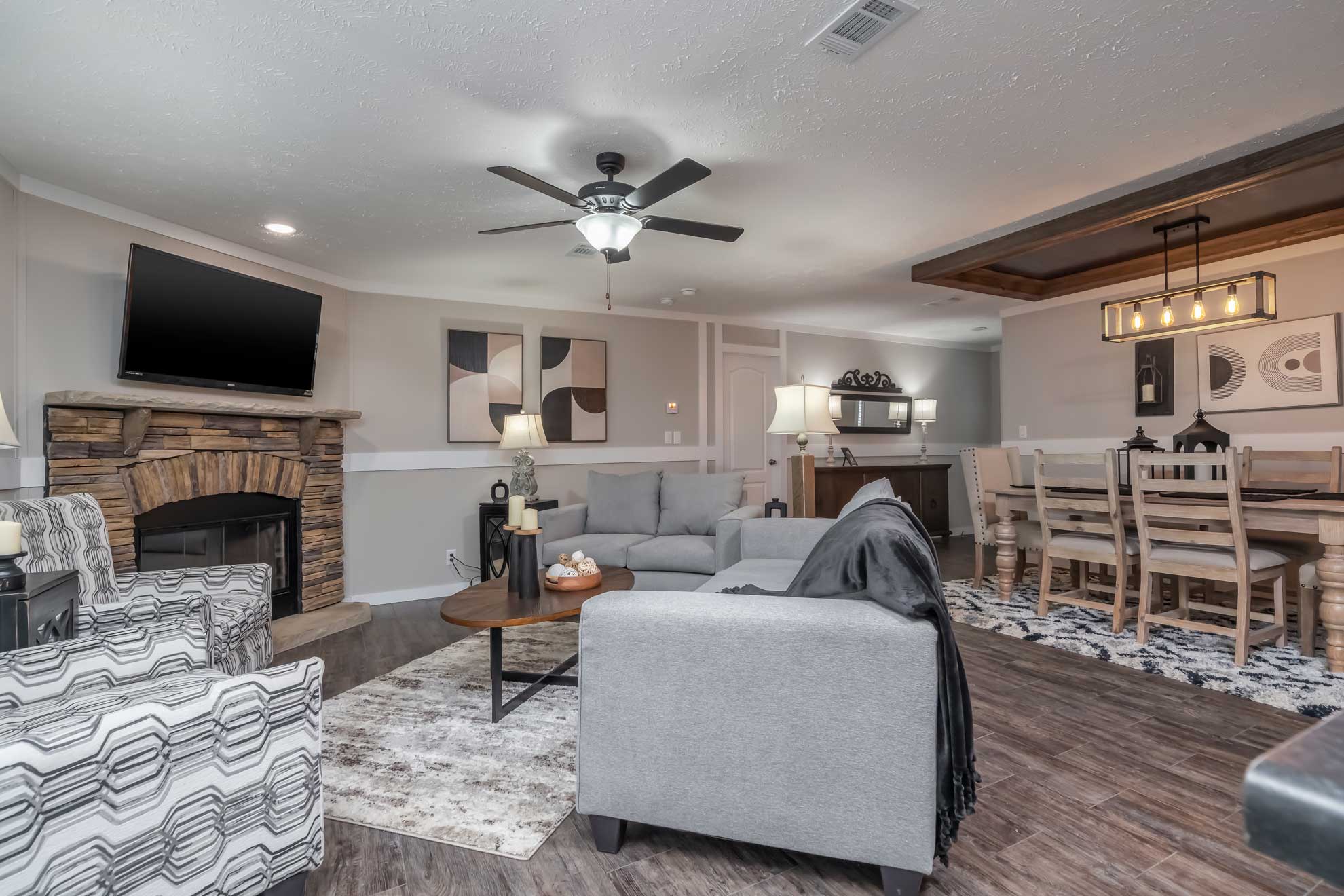 Modern living and dining area with a cozy gray sofa, patterned armchairs, and wooden coffee table. Stone fireplace, abstract art, and dining set create a warm, inviting ambiance.