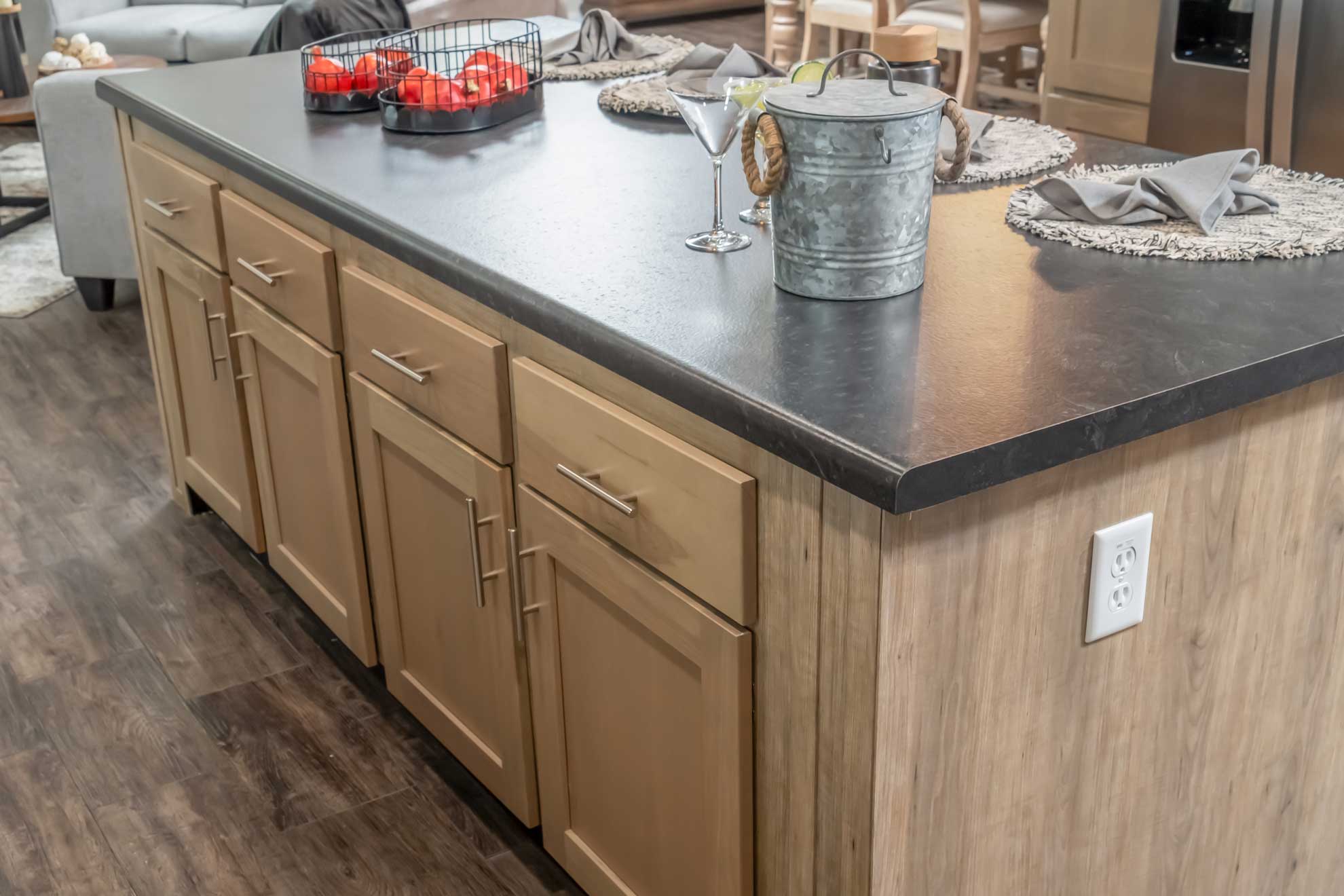 Kitchen island with a dark countertop and light wood cabinets. It holds a metal bucket, a martini glass, and a basket of red apples, creating a cozy, inviting atmosphere.
