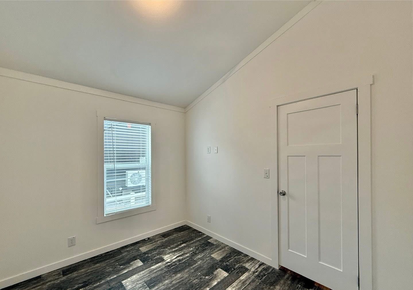 Empty room with white walls, a slanted ceiling, and dark wood-like flooring. Features a window with blinds and a closed white door, conveying a clean, minimalist look.