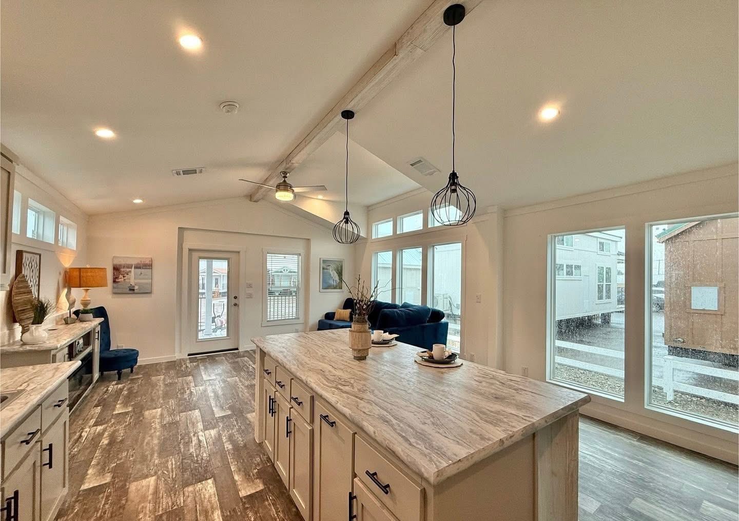 Spacious modern kitchen with a large marble island, pendant lights, wood floors, and bright windows. Cozy seating and decor create a welcoming atmosphere.