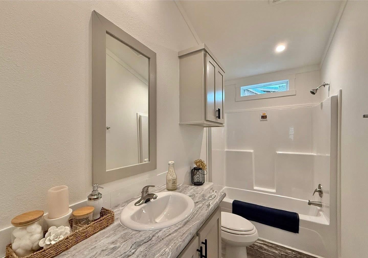 Modern bathroom with a white and gray color scheme. Features a marble countertop, stylish sink, mirror, overhead cabinets, and a bathtub with a blue towel.