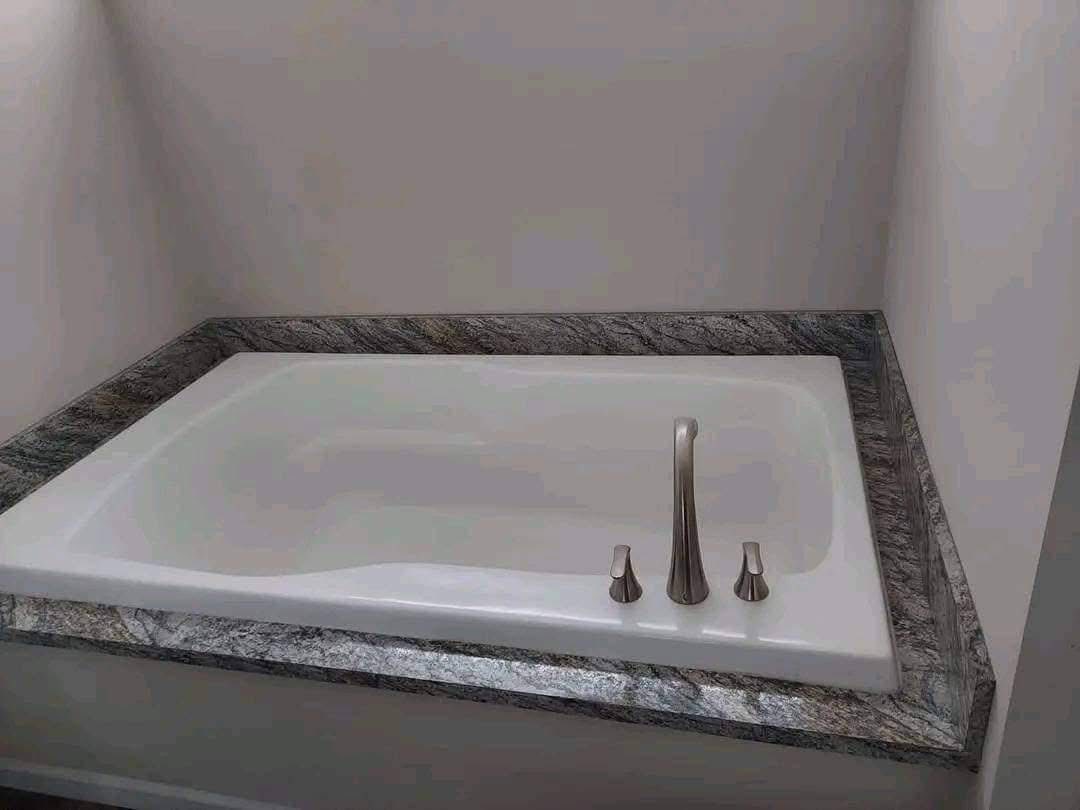 A white rectangular bathtub with silver fixtures is set in a sleek granite surround. The minimalistic design creates a calm and modern ambiance.