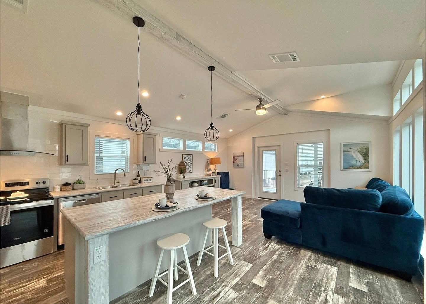 Modern open-plan kitchen and living area with light wood floors. Features an island with stools, hanging lights, dark blue sofa, and large windows. Cozy and airy ambiance.