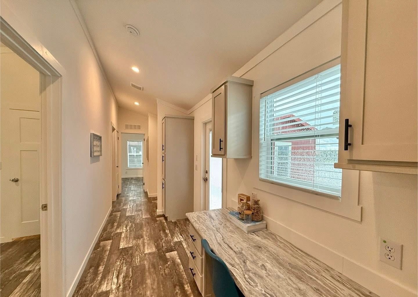 A bright, narrow hallway with a wood-textured floor, cream-colored walls, and recessed lighting. A window and countertop with cabinets are on the right.