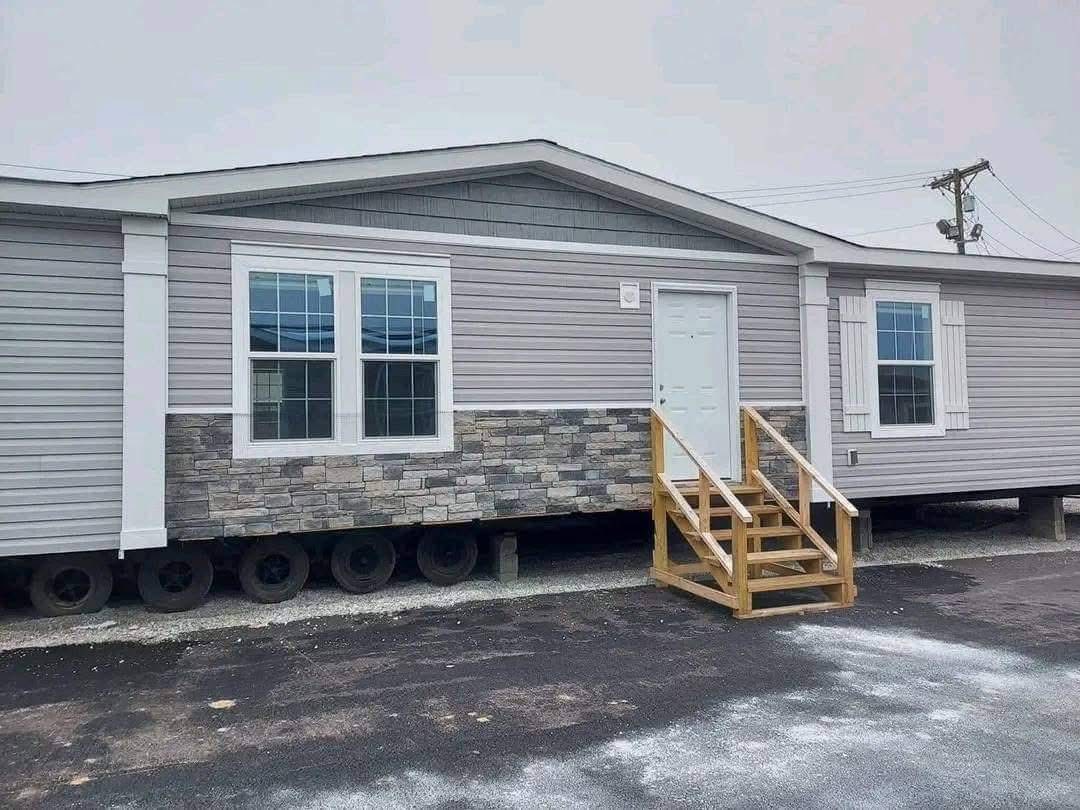 A light gray mobile home with a stone facade and white trim. It features two large windows and a white door, accessed by wooden steps. Overcast sky.