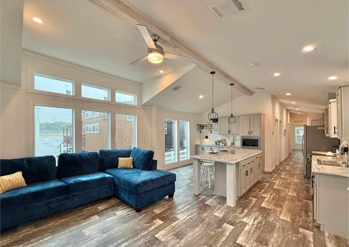 Spacious open-concept living area with blue sectional sofa, vaulted ceilings, large windows, and a kitchen with an island, emitting a cozy, modern vibe.