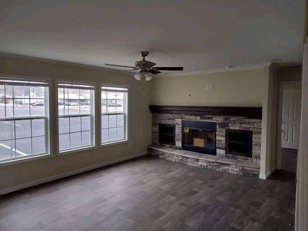 A spacious living room with large windows, a ceiling fan, and a stone fireplace. The room has dark wood flooring and neutral-colored walls.