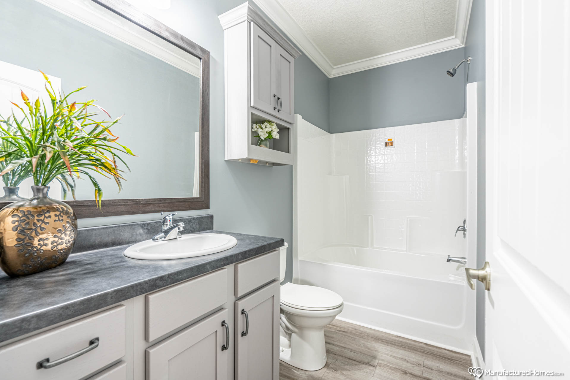 Modern bathroom with gray walls and wood flooring. Features a white vanity with a dark counter, sink, large mirror, and a potted plant. Bathtub and toilet in the background.