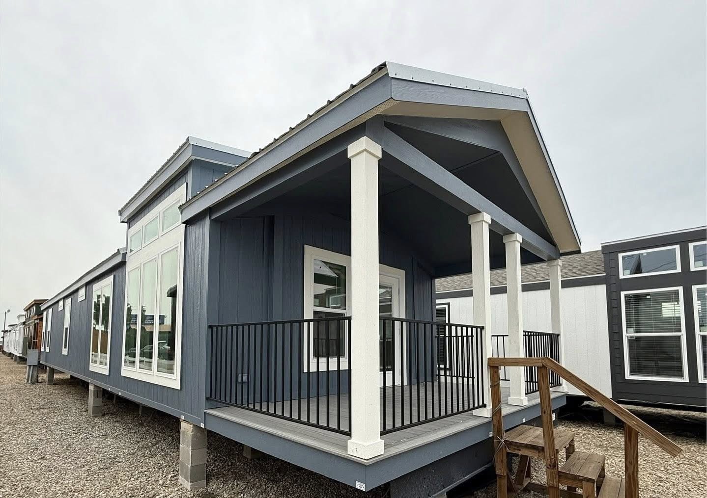 Blue modular home with a gabled roof and large windows. It features a small porch with white columns and black railing, set on gravelly ground.