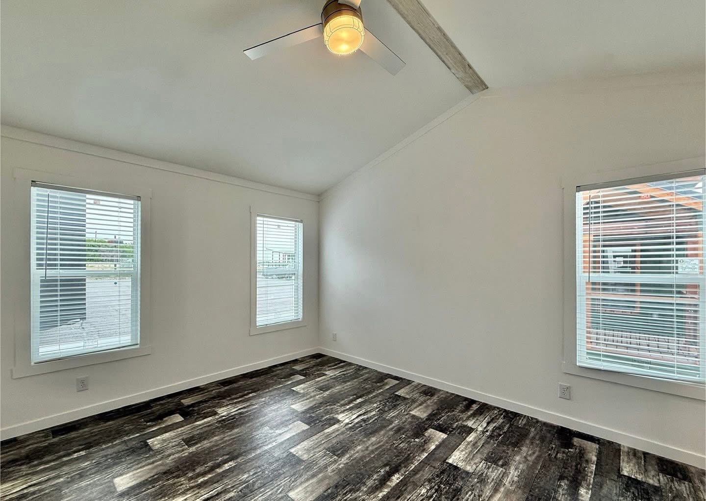 Minimalist room with white walls, dark wood flooring, and three large windows letting in natural light. Ceiling fan centered above adds a modern touch.