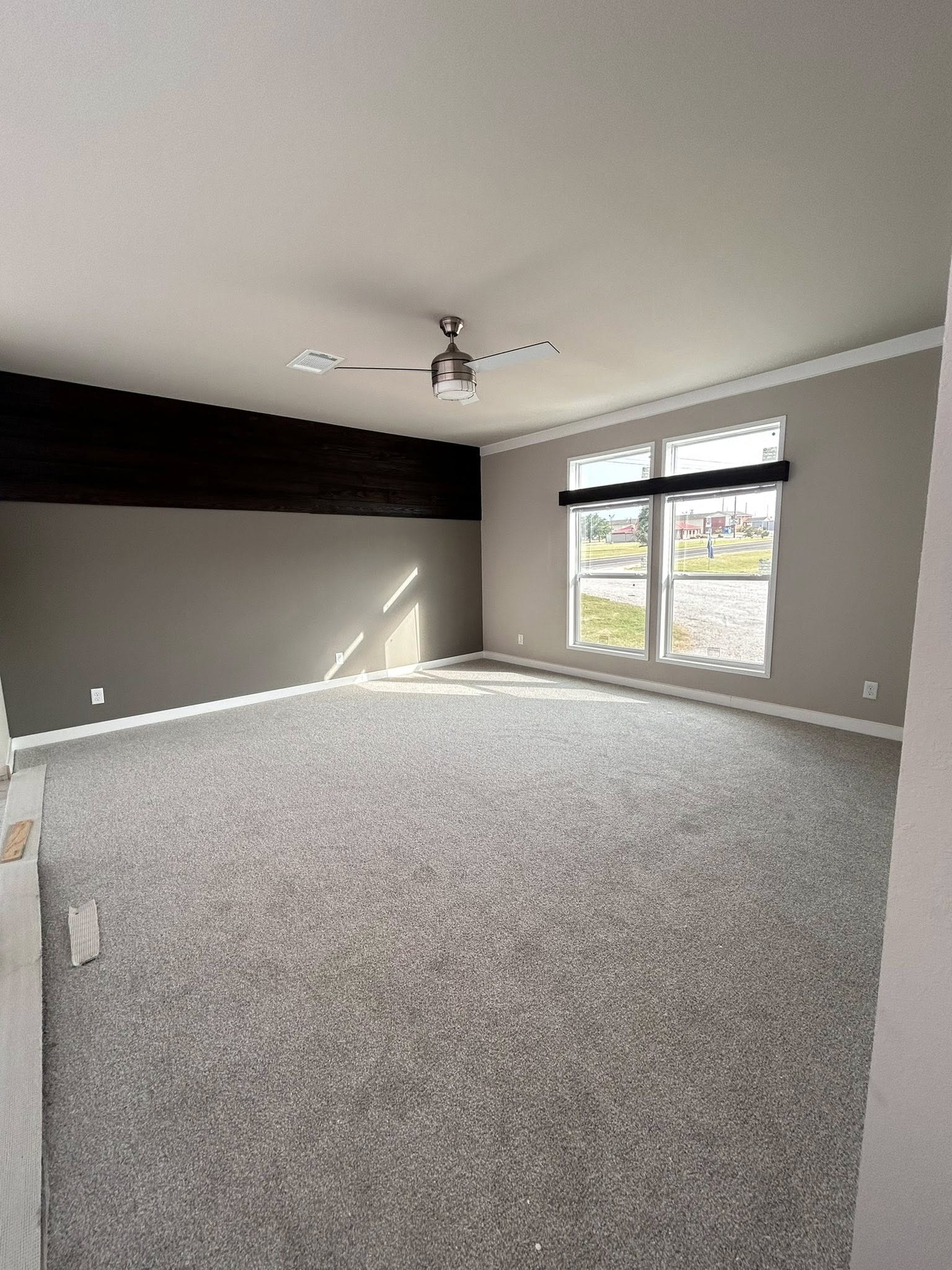 Empty room with gray carpet and beige walls, featuring a dark wood accent wall. Ceiling fan in the center, large windows with sunlight streaming in. Cozy and modern.
