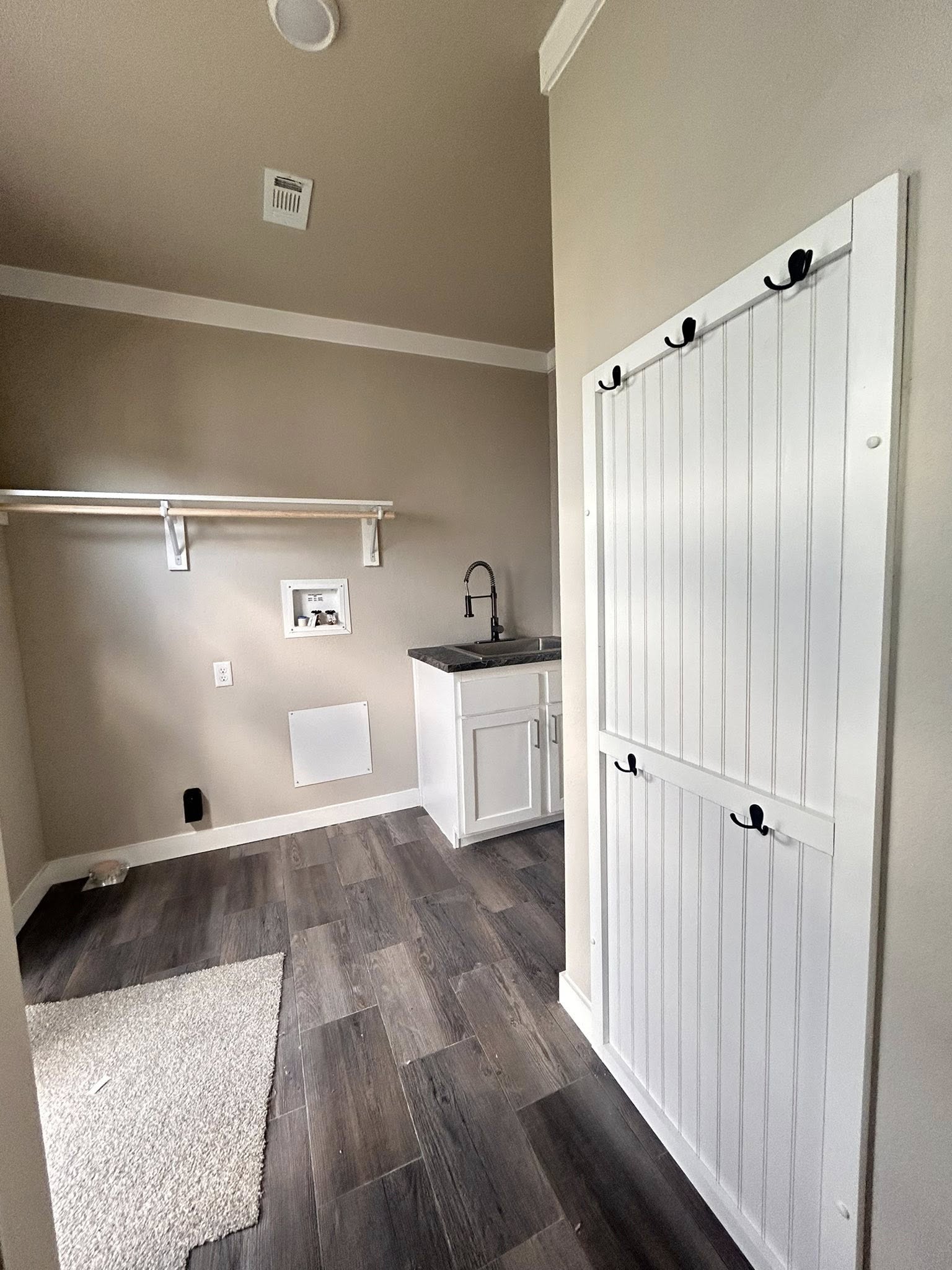 Laundry room with wood flooring, beige walls, and white trim. Features a white cabinet with a sink, wall-mounted shelf, and hooks on the door.