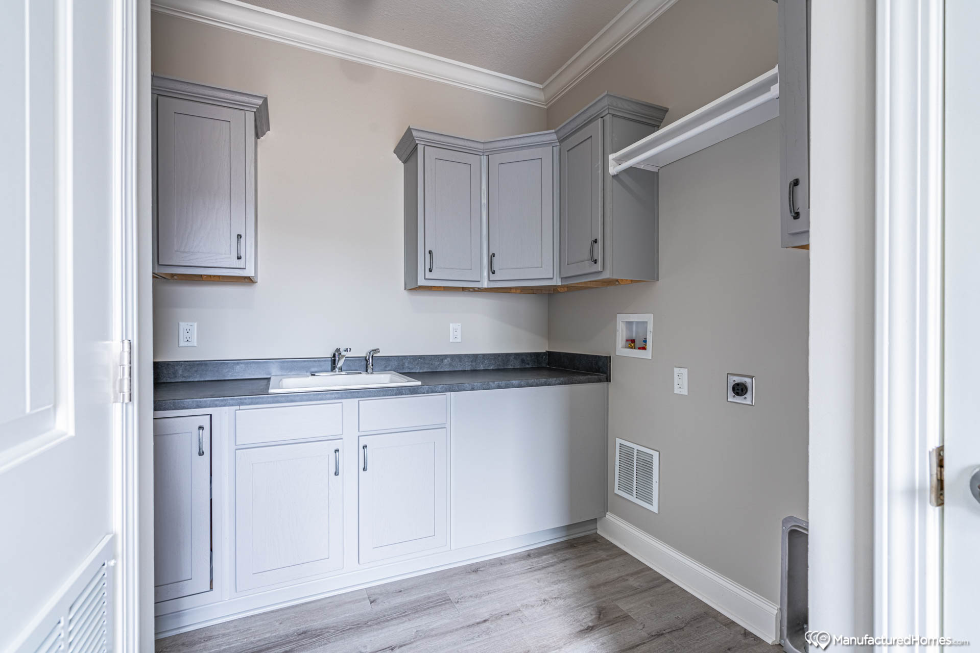 Laundry room with light gray cabinets, a sink, and dark countertop. Beige walls, white trim, and wood flooring create a clean, modern look.