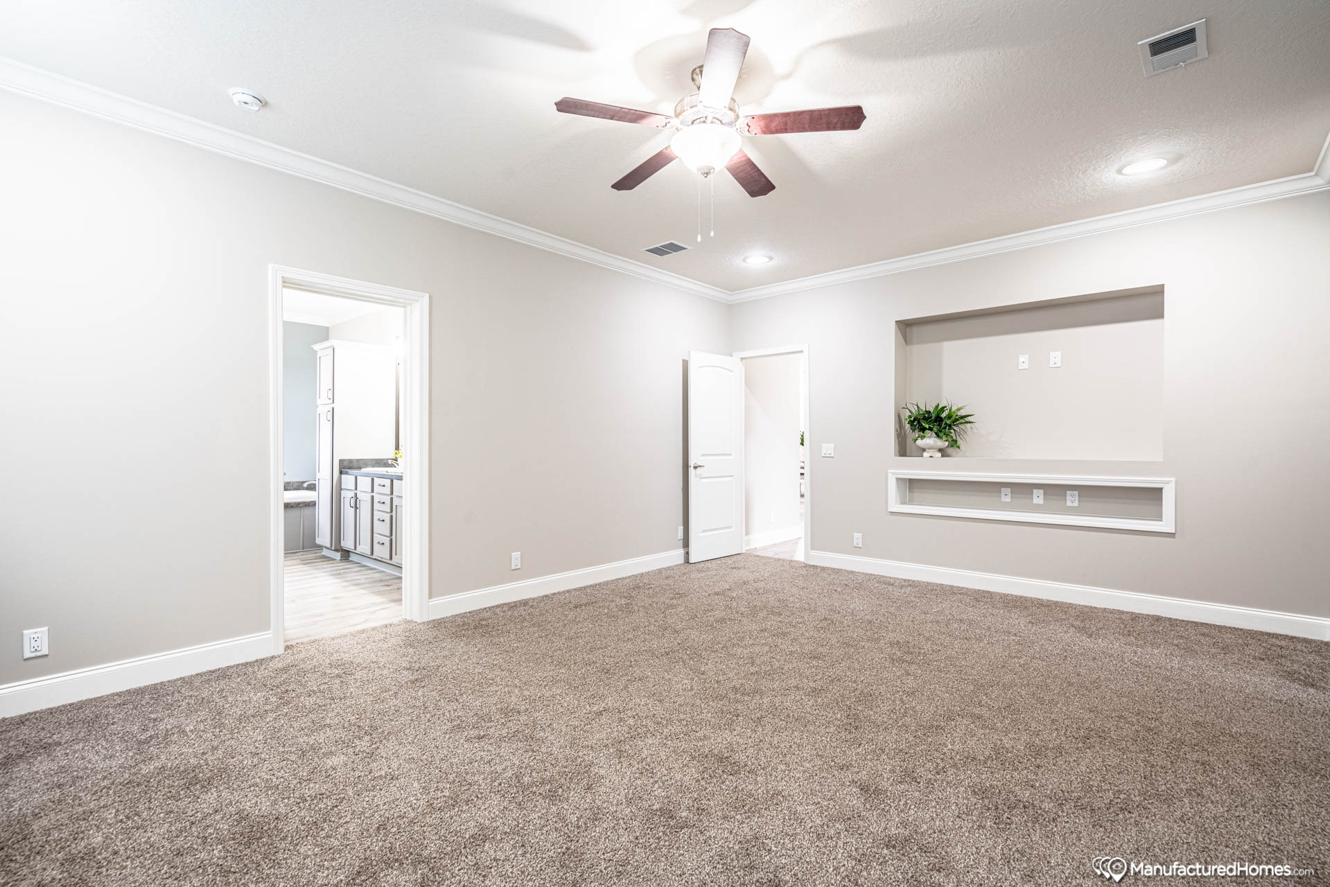 Spacious carpeted room with neutral walls, ceiling fan, and recessed lighting. Features include an open doorway to a bright bathroom and a built-in shelf with a plant.