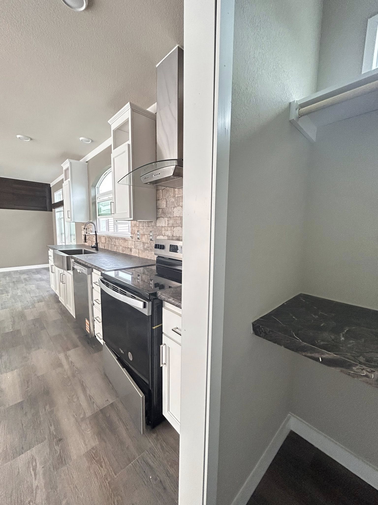 Modern kitchen with light wood cabinets, black appliances, and stone backsplash. Adjacent is a small closet with a dark marble shelf, conveying a sleek, minimalist tone.