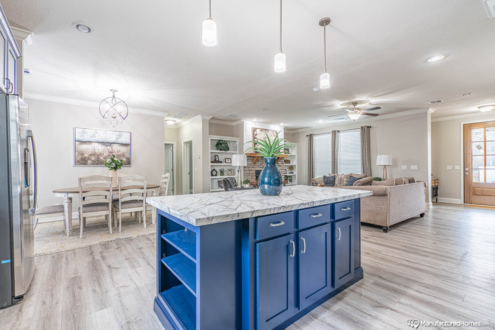 Spacious open-plan kitchen and living area. Features a blue island with marble top, pendant lights, dining table, beige sofas, and wood floors. Inviting and modern.
