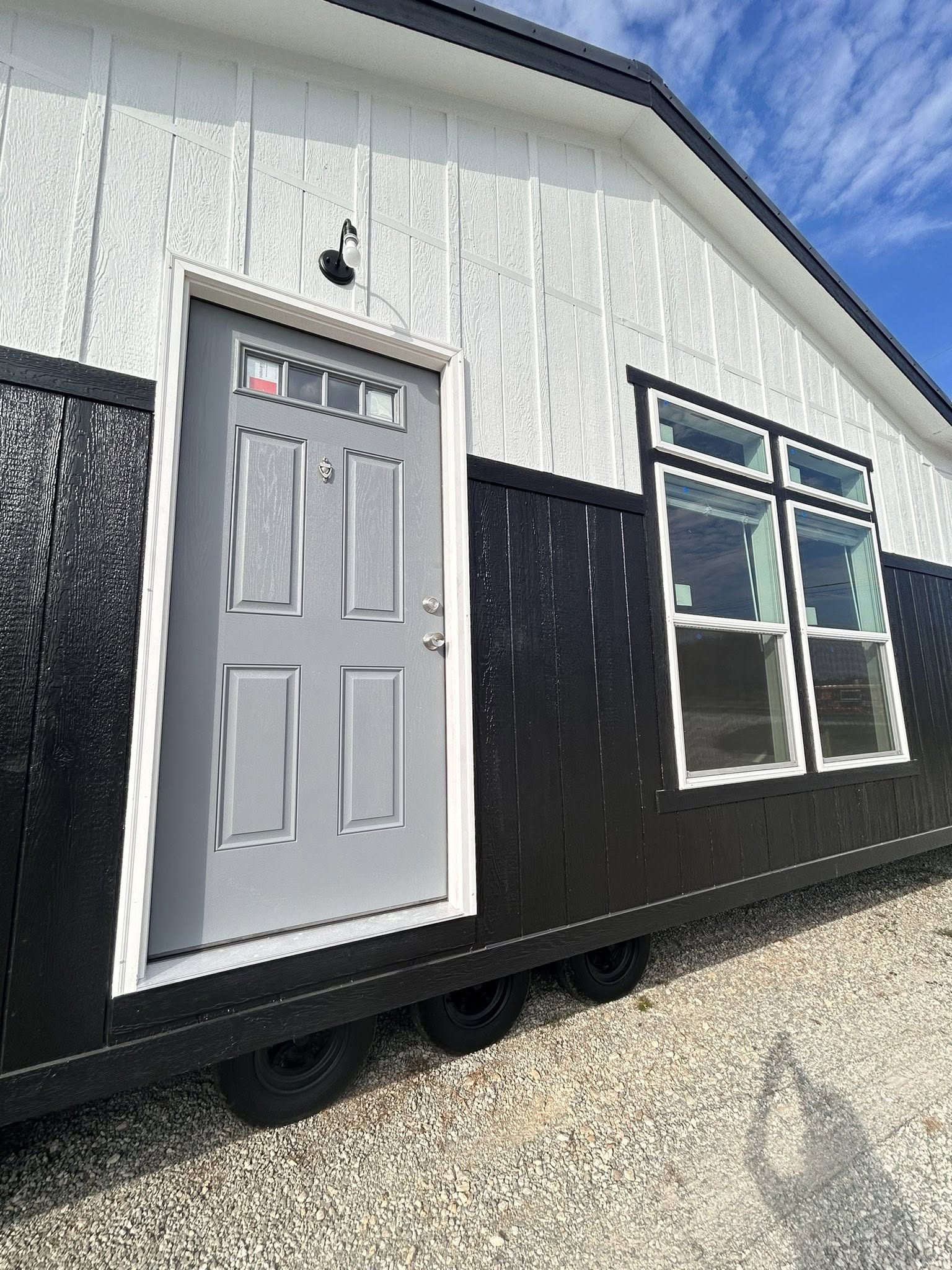 Small, modern black-and-white tiny house with a gray door and large windows. The house rests on wheels, set against a bright blue sky.